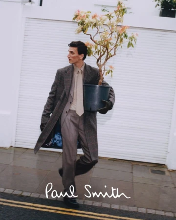 A man in a grey suit and coat carries a potted plant in front of a white garage door, showcasing a blend of fashion and nature.