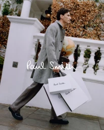 A man in a grey wool coat walking past white neoclassical columns carrying shopping bags and a globe