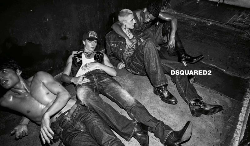 Four male models in denim jeans and leather boots recline on a concrete floor in a black and white fashion campaign image.