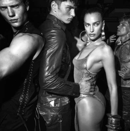 Black and white fashion shot featuring a woman in a bodysuit flanked by three men, against a dark background.