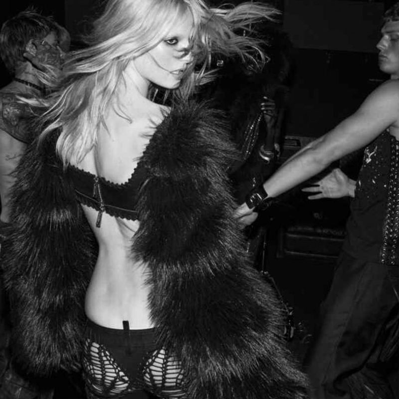Monochrome fashion shot of a woman in a fur coat and lingerie standing in a dark environment, with other people in the background.