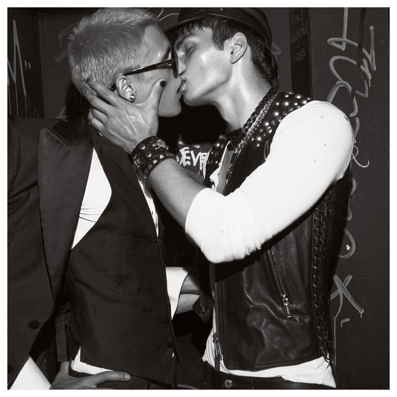 Monochrome portrait captures two men kissing, one in a suit and glasses, the other in a studded leather vest and cap, against graffiti wall.