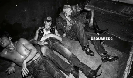 Four male models in denim jeans and leather boots recline on a concrete floor in a black and white fashion campaign image.