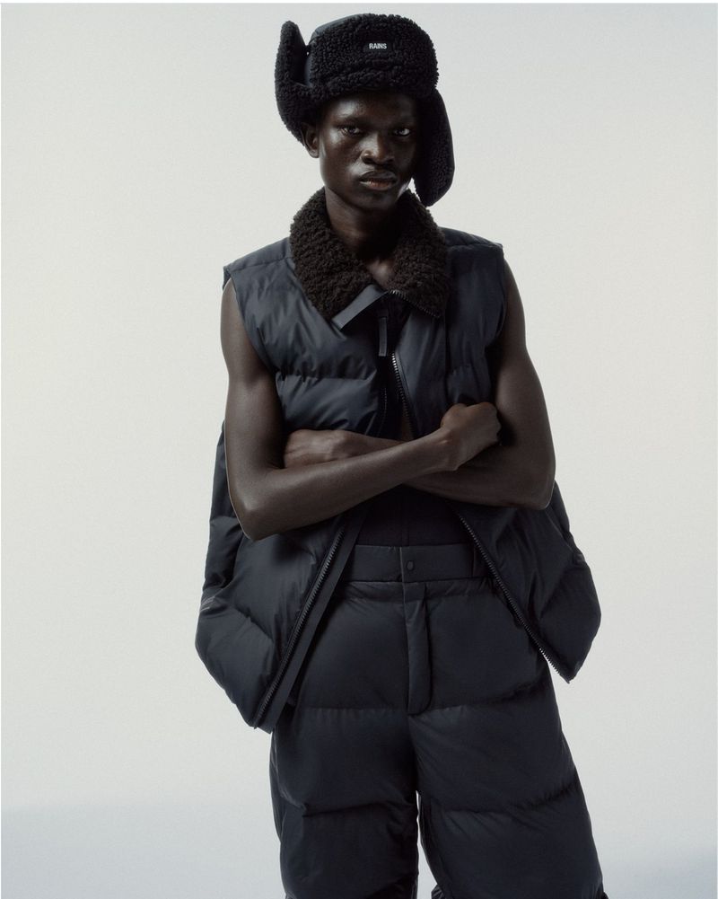 Fashion shot of a dark-skinned male model in a black trapper hat and black quilted vest and pants against a plain white background.