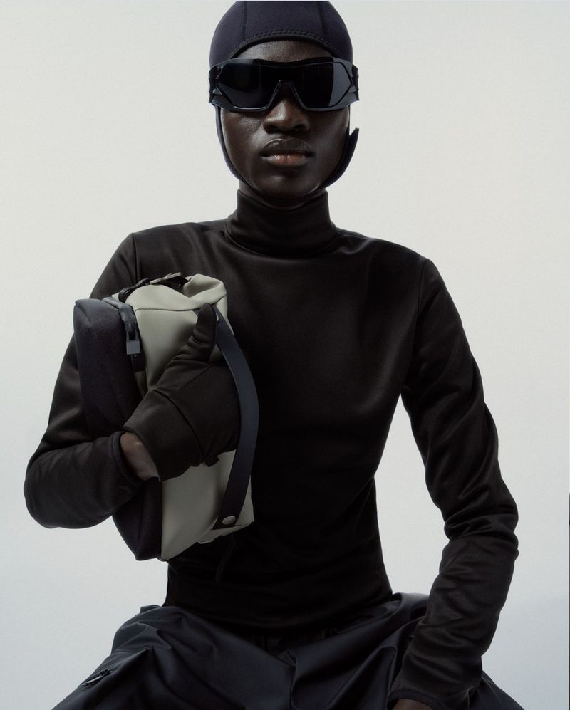 Dark-skinned model in futuristic black head covering and sunglasses, holding a stylish two-tone gray handbag on a neutral background.