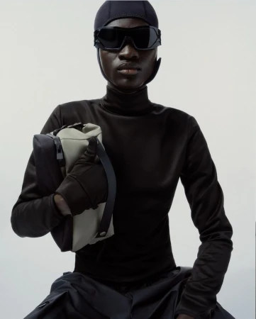 Dark-skinned model in futuristic black head covering and sunglasses, holding a stylish two-tone gray handbag on a neutral background.