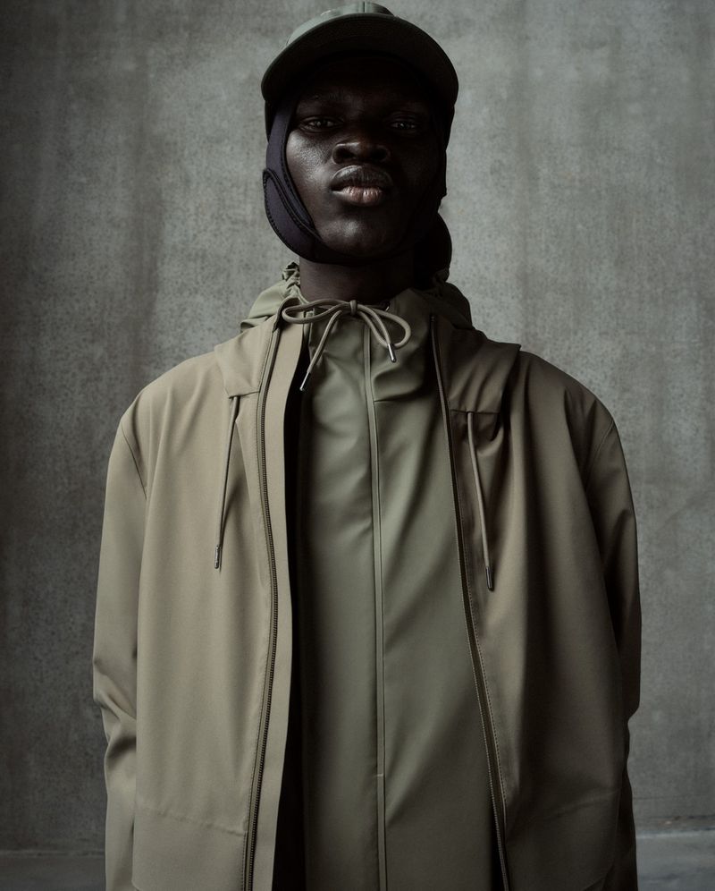 A stylish dark-skinned male model wears a muted green coat and hat against a grey backdrop, showcasing contemporary fashion.