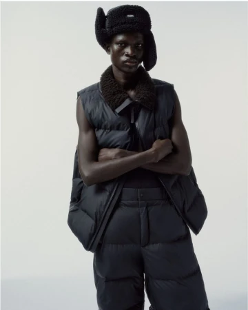 Model Chubath Kutien wearing a black sleeveless puffer vest and trapper hat against a white studio backdrop for Rains F/W 2025.