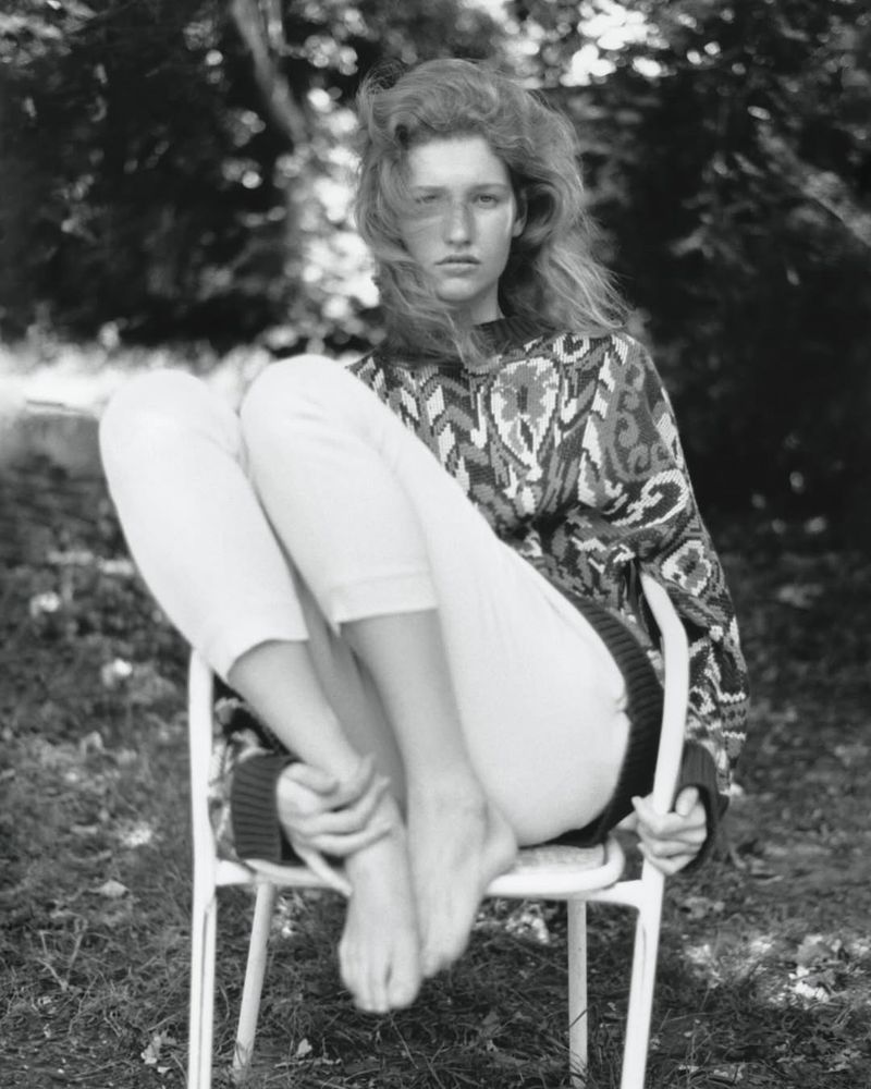 Young woman with long hair poses seated on a chair outdoors, wearing a patterned sweater and white pants in a monochrome photograph.