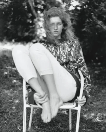 Young woman with long hair poses seated on a chair outdoors, wearing a patterned sweater and white pants in a monochrome photograph.