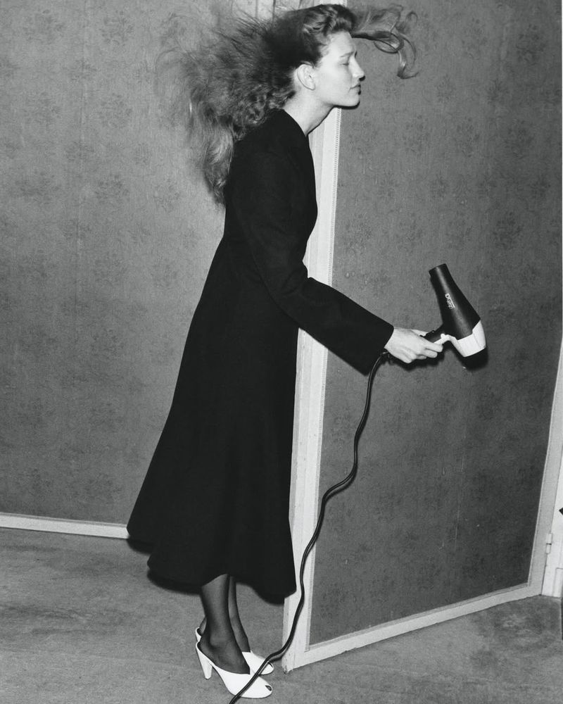 A woman in a black coat and white shoes stands sideways holding a hair dryer, her hair blowing. Against a grey background.