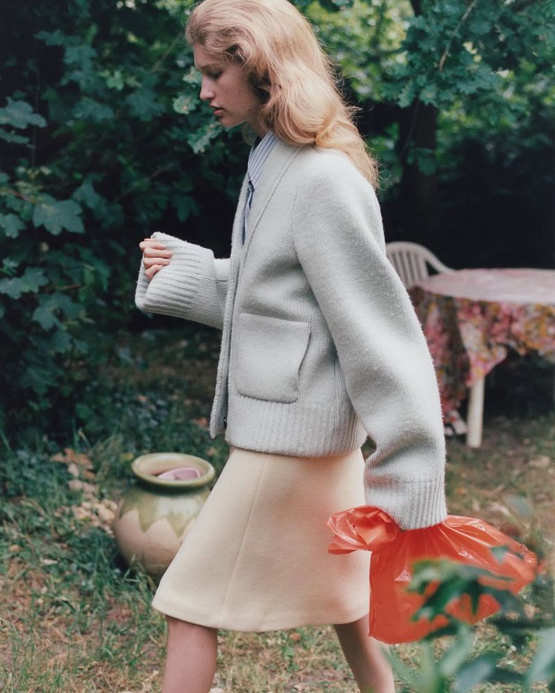 A young woman in a cardigan and skirt walks through a garden carrying a plastic bag with foliage and a table visible in the background.
