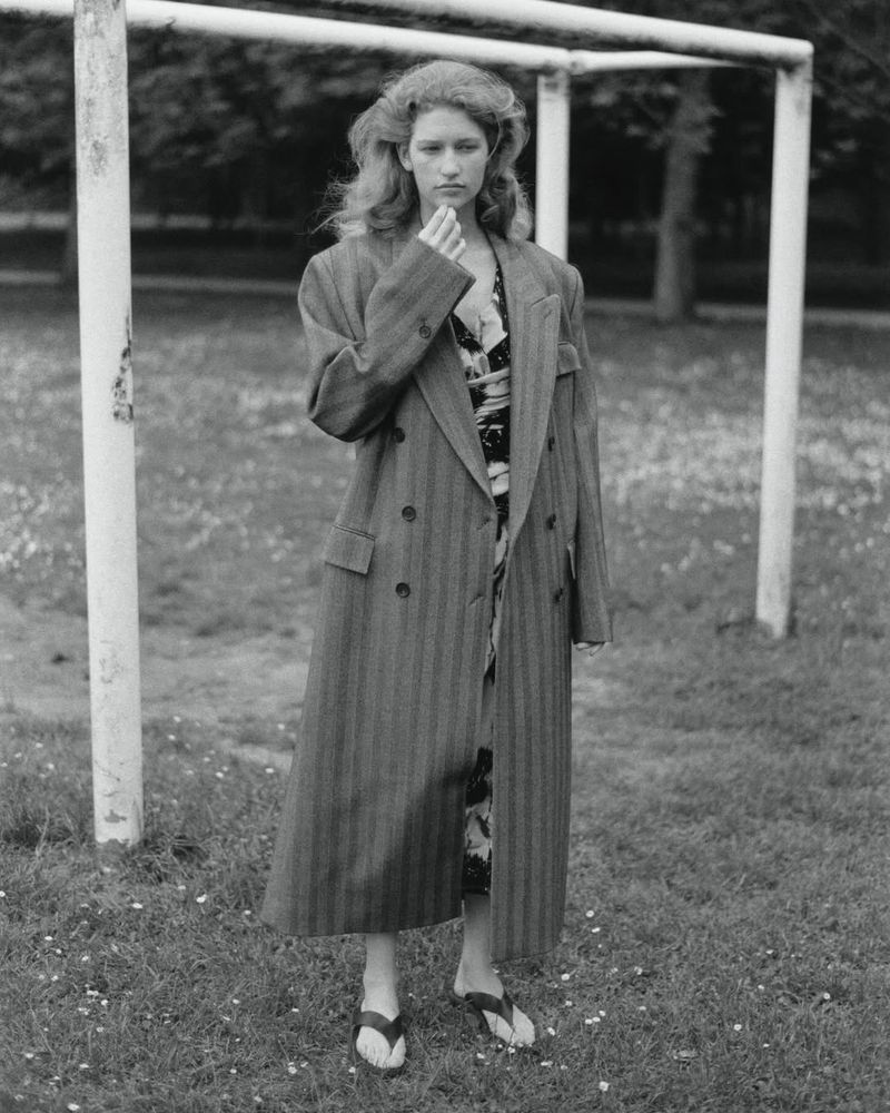 Black and white portrait of a woman standing in a striped coat on a grassy field near white goalposts, with trees in the background.