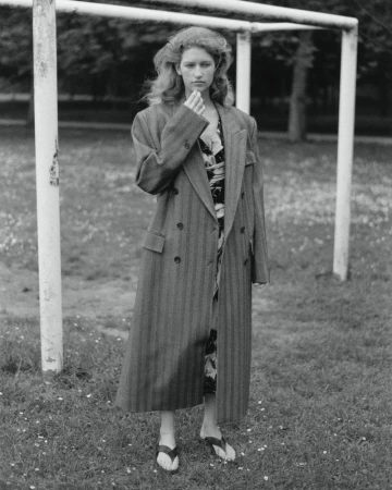 Black and white portrait of a woman standing in a striped coat on a grassy field near white goalposts, with trees in the background.