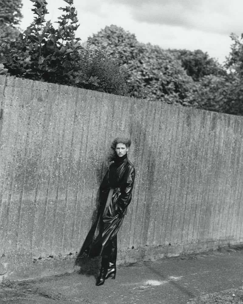 Model Apolline Rocco Fohrer posing in a long dark belted trench coat against a textured vertical wall outdoors.