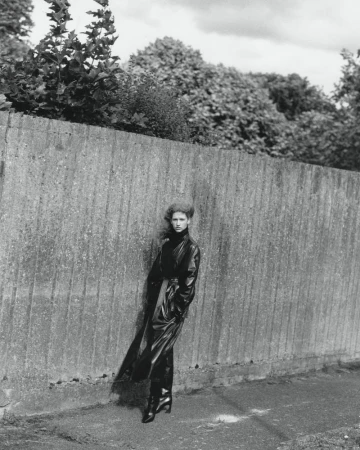 Model Apolline Rocco Fohrer posing in a long dark belted trench coat against a textured vertical wall outdoors.