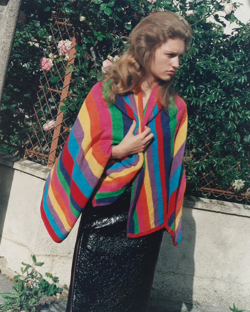 A young woman with blond hair stands outdoors wearing a rainbow-striped coat and a shiny black skirt, posing in front of plants and a fence.