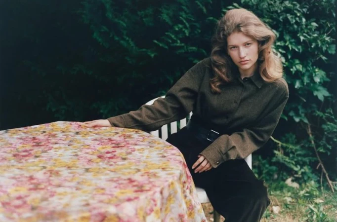 Model Apolline Rocco Fohrer wearing an olive button-down shirt and black trousers, seated outdoors beside a floral-draped table.
