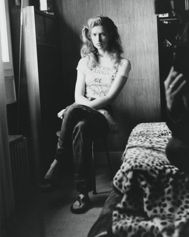 Black and white photo of a fair-skinned, blonde woman sitting on a chair indoors, wearing a graphic tee and pants, with shadows.