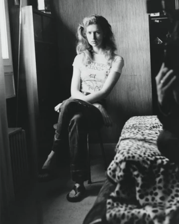 Black and white photo of a fair-skinned, blonde woman sitting on a chair indoors, wearing a graphic tee and pants, with shadows.