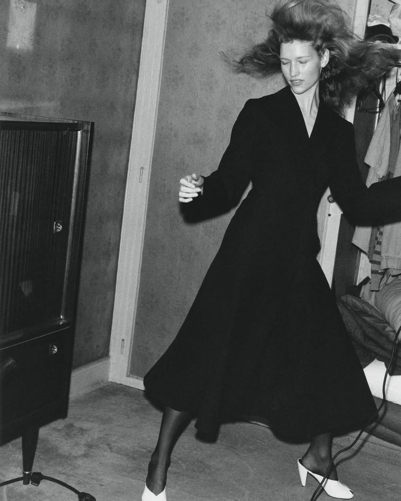 A woman dances in a monochrome photo wearing a dark dress and light heels, with an old radio in a room, capturing a retro fashion vibe.