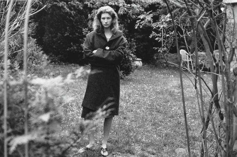 A young adult woman in a dark coat stands with crossed arms in a monochrome outdoor portrait with trees and grass.
