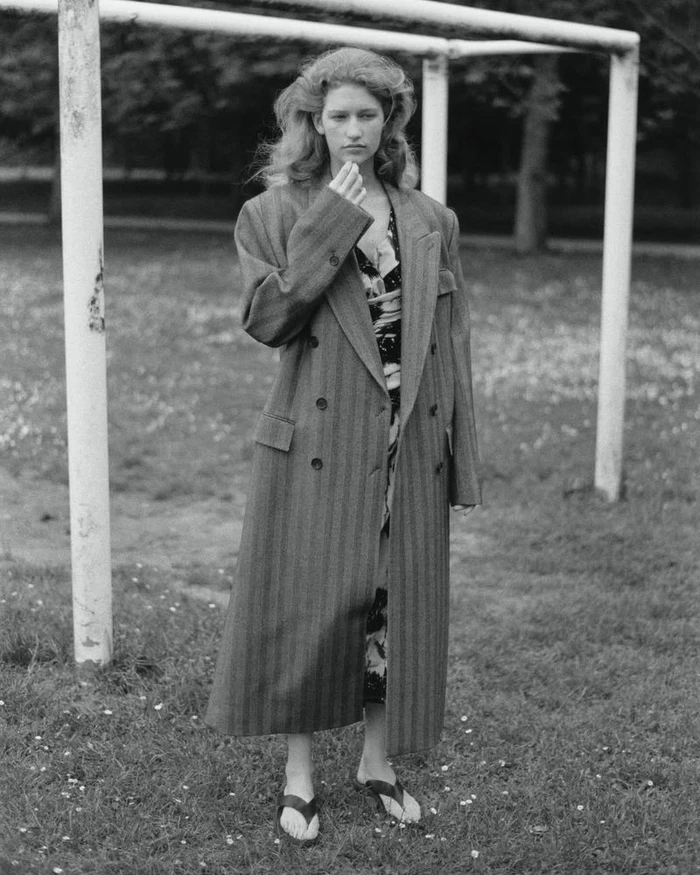 Model Apolline Rocco Fohrer poses in an oversized tailored coat and heeled sandals under white goalposts on a grassy field.