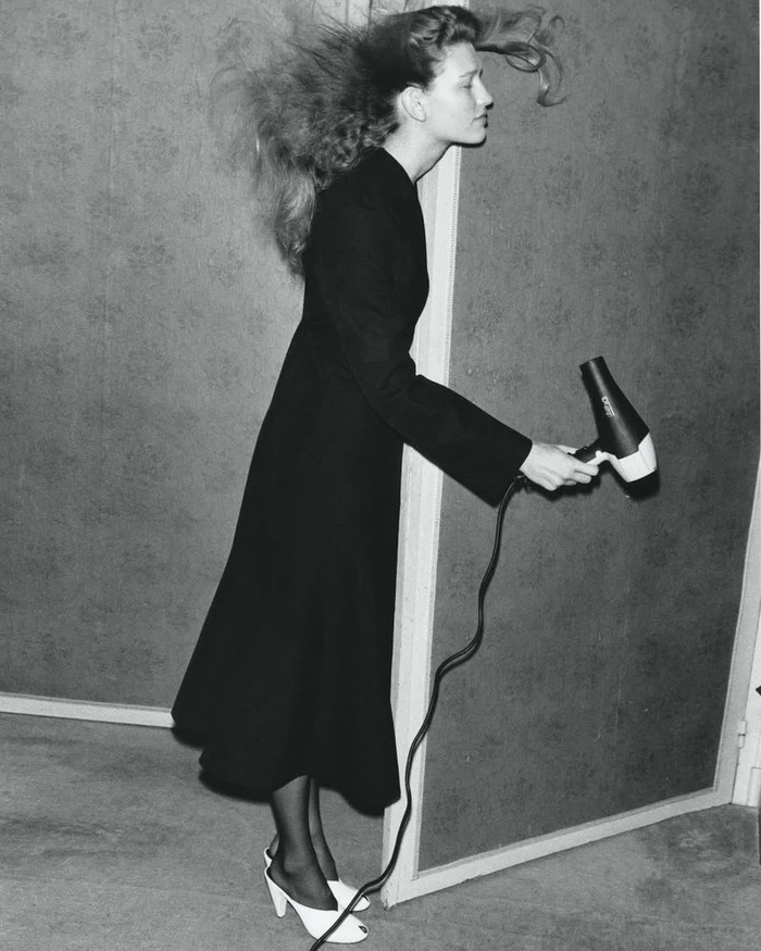 Model in a black coat holding a hairdryer with blowing hair against a patterned wall, shot by Krisztian Eder for D Repubblica.