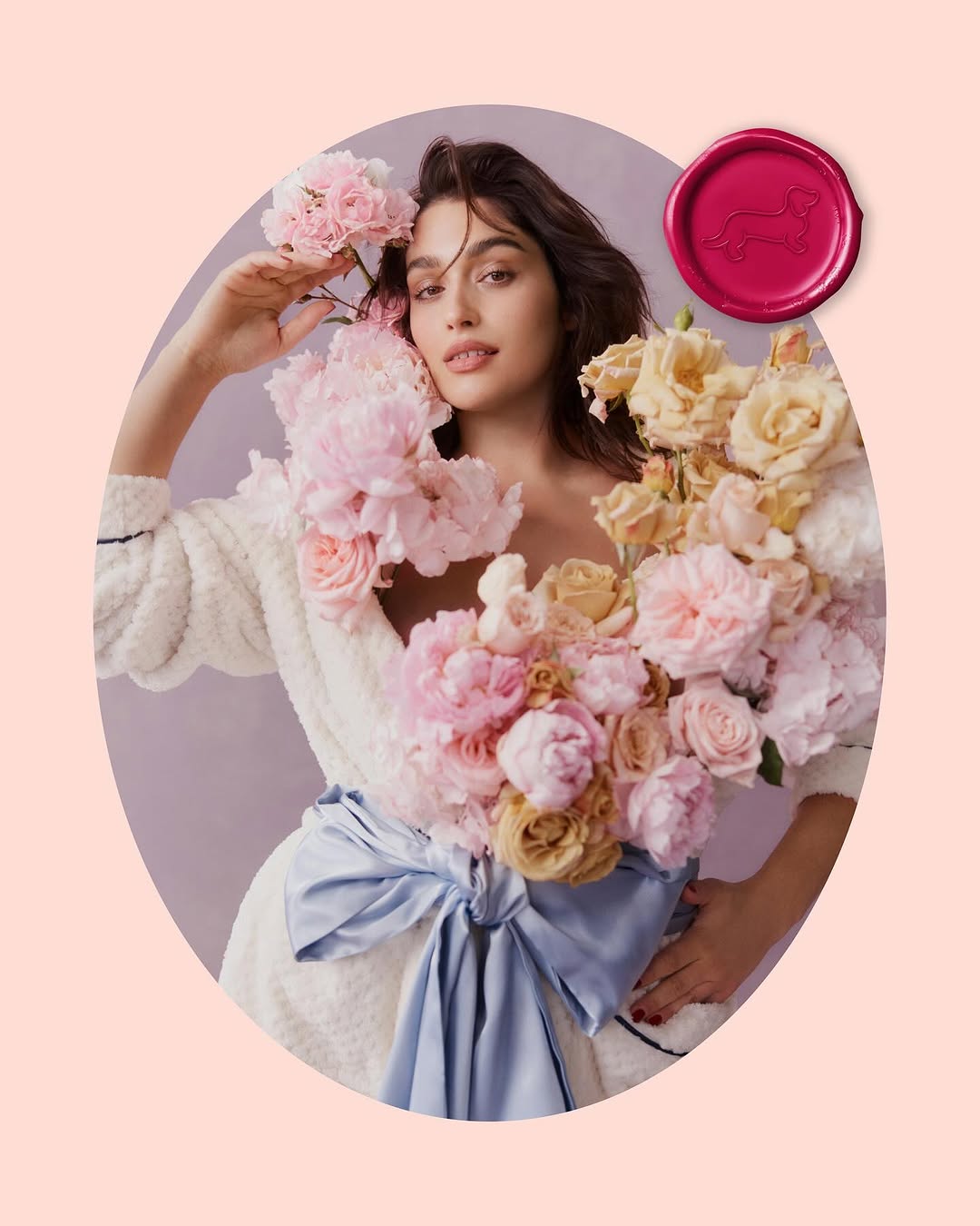 Portrait of a woman in a white robe with light brown hair, wearing a pastel flower arrangement and a light blue sash in a pink space.