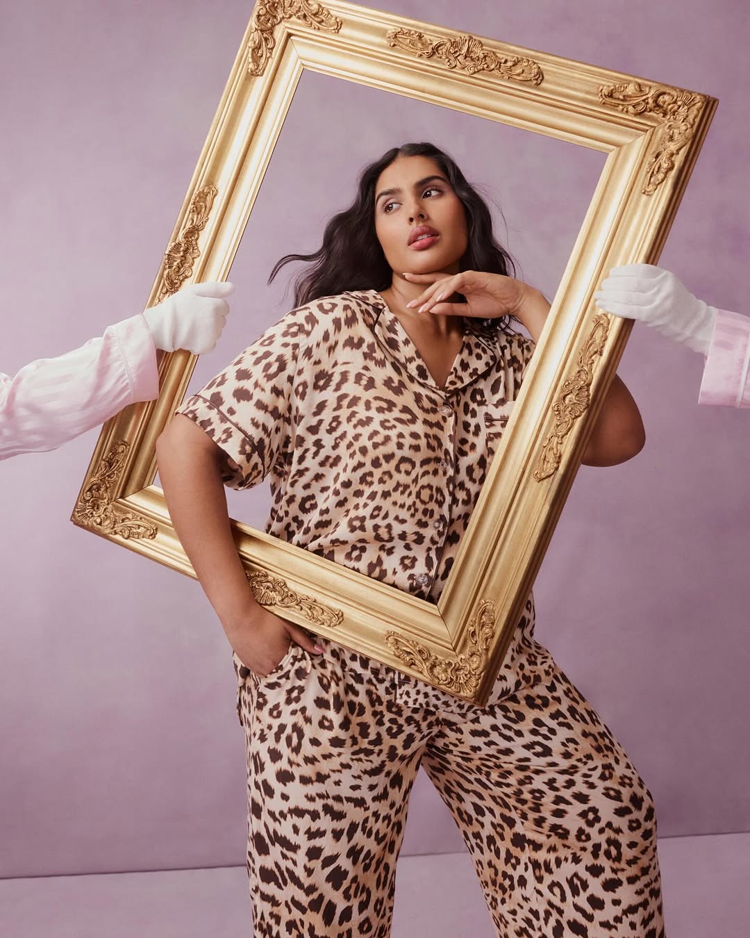 A brunette woman in leopard-print pajamas poses in a gold frame held by gloved hands against a light purple backdrop.