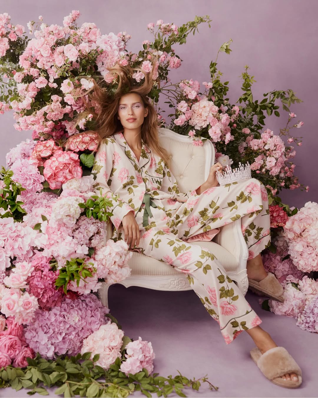 Woman in floral pajamas seated in an ornate chair surrounded by pink flowers and greenery in a studio setting with soft lighting.