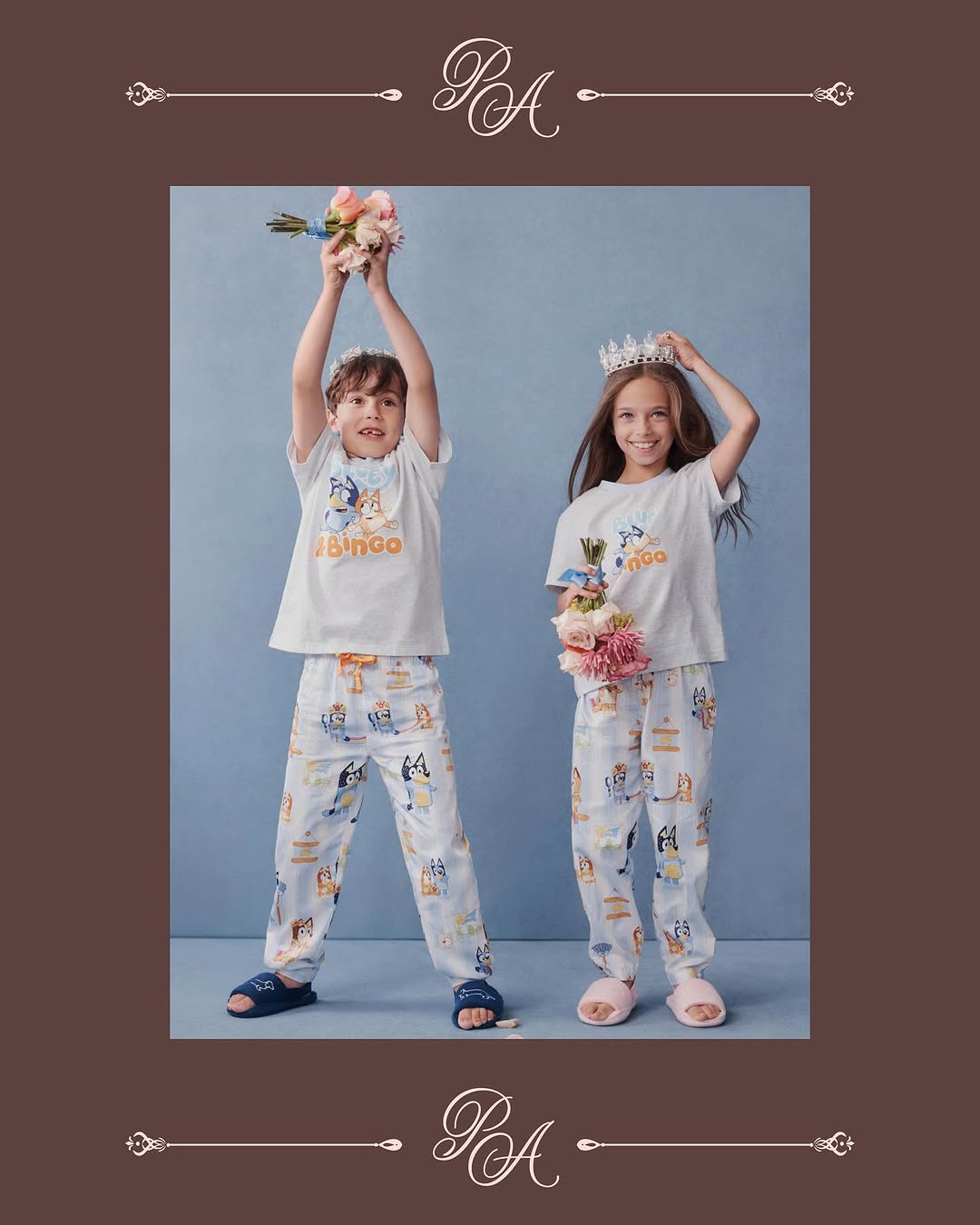 Two children in pajamas holding flowers and wearing crowns pose in front of a blue background in a studio shot. They are smiling.