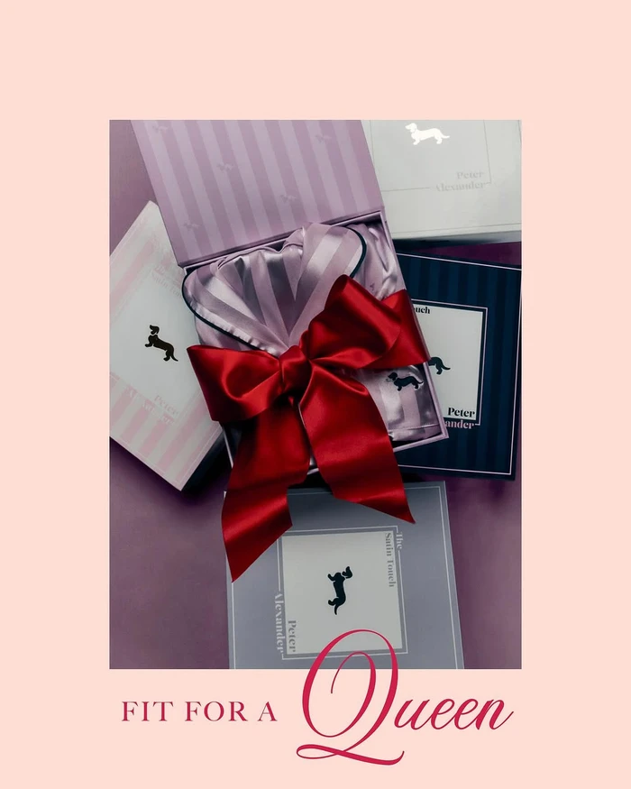 An open gift box containing striped silk pyjamas tied with a large red satin bow, surrounded by stacked branded boxes on a solid backdrop.