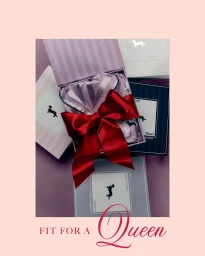 An open gift box containing striped silk pyjamas tied with a large red satin bow, surrounded by stacked branded boxes on a solid backdrop.