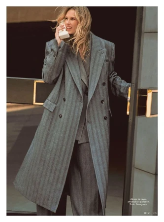 A blonde-haired woman in a gray pinstripe suit and long coat holds a vintage telephone in a fashion editorial shot indoors.