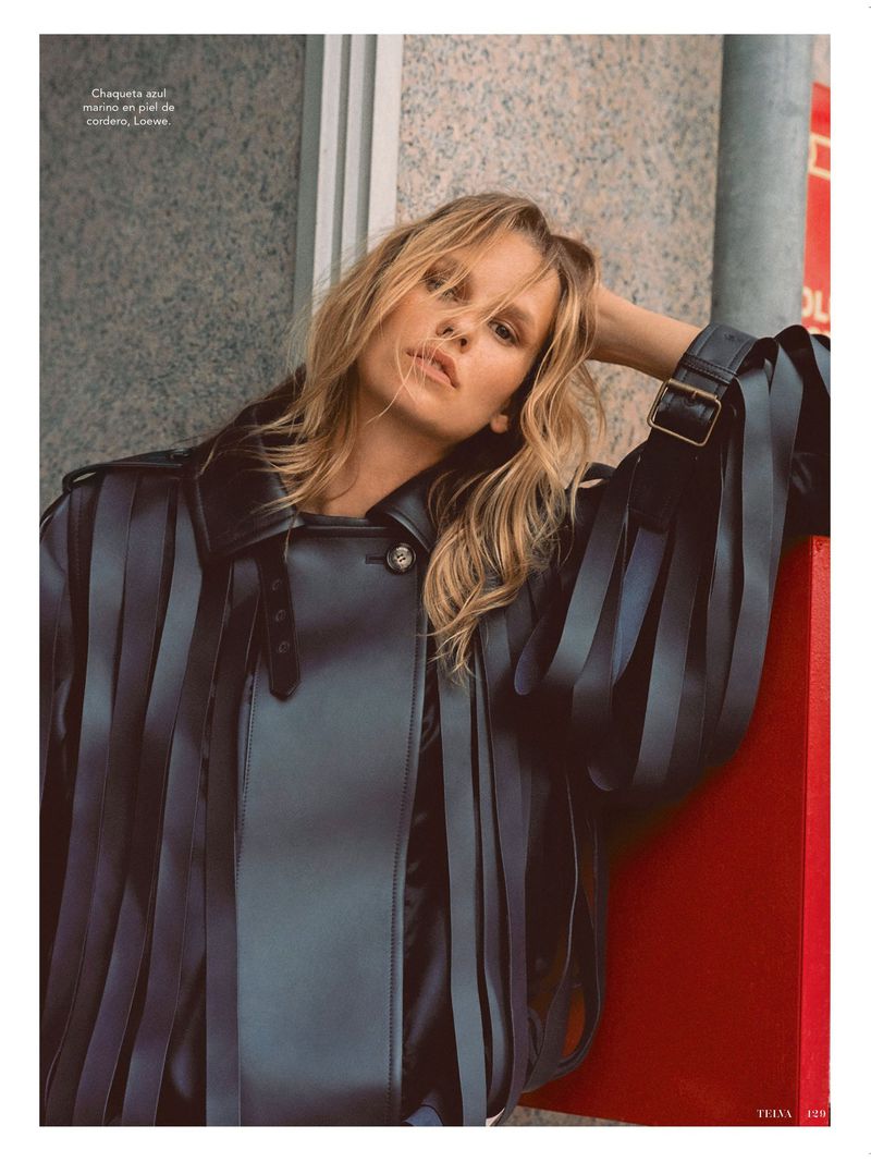 Blonde haired woman poses in leather jacket with fringe detail in an urban setting, shot in medium portrait orientation with neutral expression.