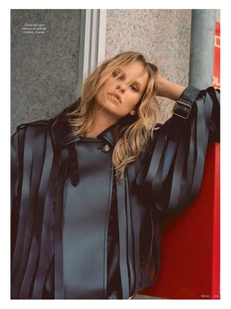 Blonde haired woman poses in leather jacket with fringe detail in an urban setting, shot in medium portrait orientation with neutral expression.