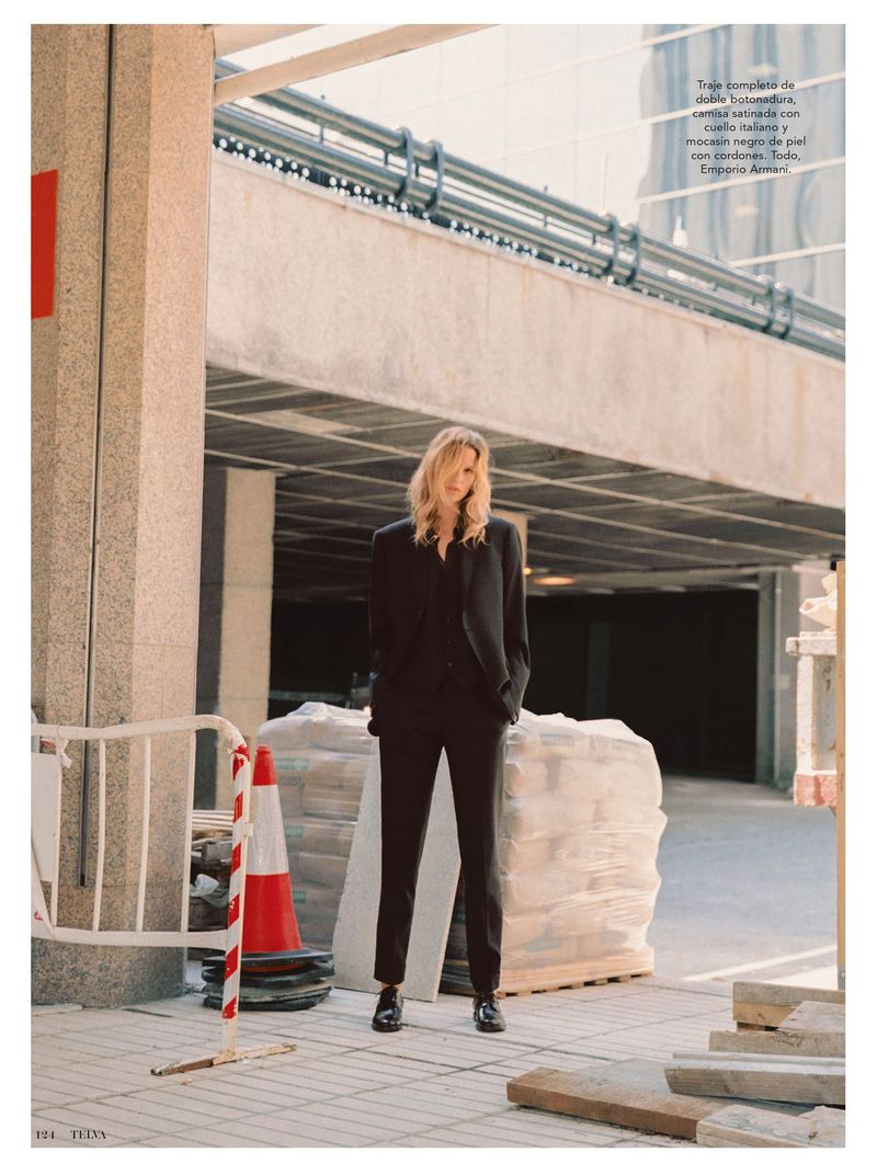 A tall blonde woman in a black suit poses on a city street, surrounded by construction materials, with a large commercial building behind.