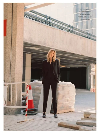 A tall blonde woman in a black suit poses on a city street, surrounded by construction materials, with a large commercial building behind.