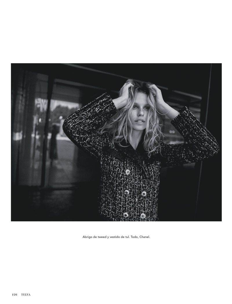 Black and white portrait of a blonde adult woman wearing a patterned jacket, gazing at the camera with hands touching her hair.