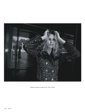 Black and white portrait of a blonde adult woman wearing a patterned jacket, gazing at the camera with hands touching her hair.