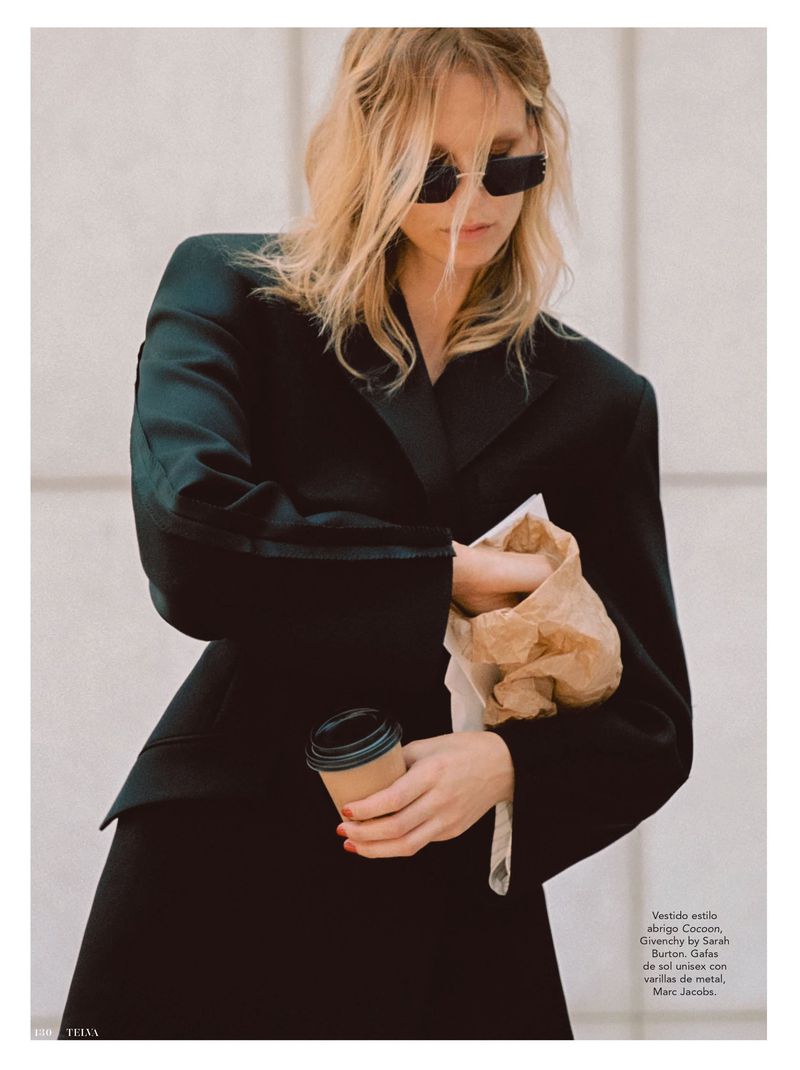 A blonde woman in a black suit holds a coffee cup and a takeout bag in a bright, overcast outdoor setting, with a fashion editorial style.