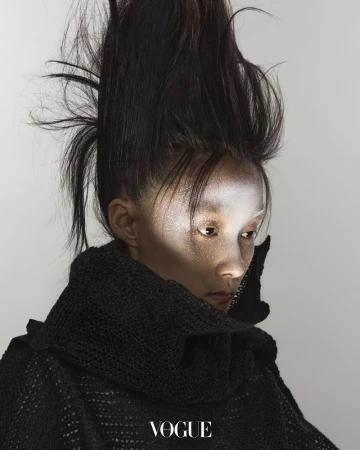 Close-up portrait of a person with dramatic hair and makeup, wearing a black knit garment, against a light neutral background. Vogue logo visible.