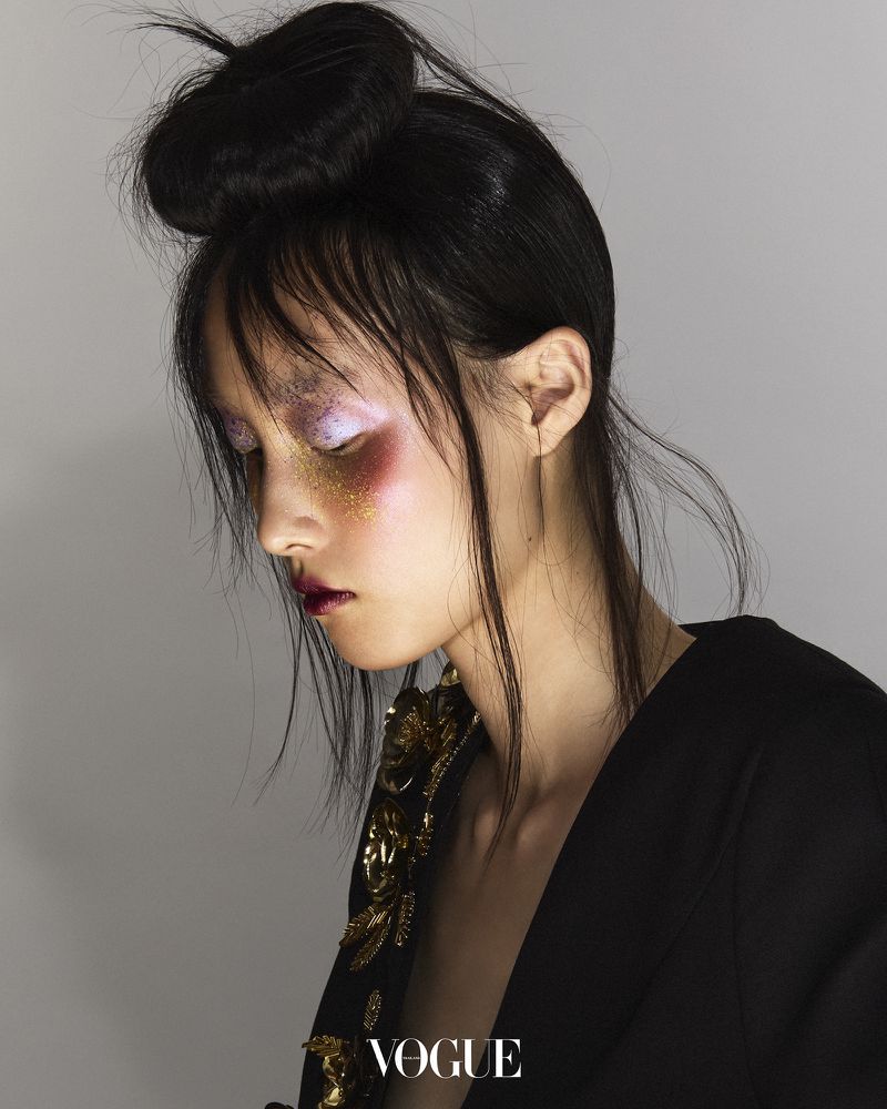 Profile of a young woman with creative makeup and dark hair styled in a bun, wearing a black jacket, against a neutral gray background.