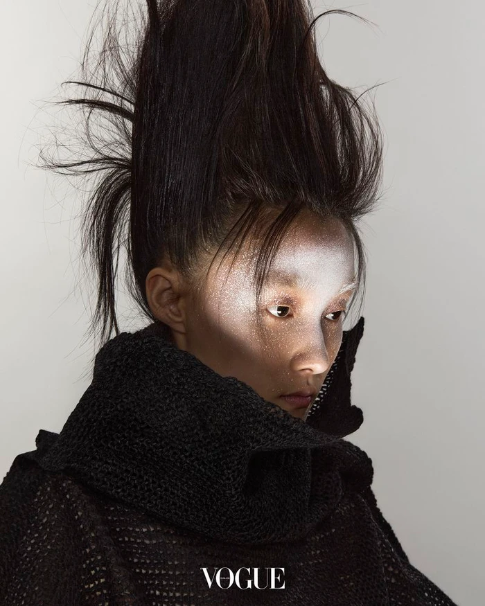 Model with avant-garde sculpted high hair and silver face makeup wearing a black textured knit top against a grey studio background.