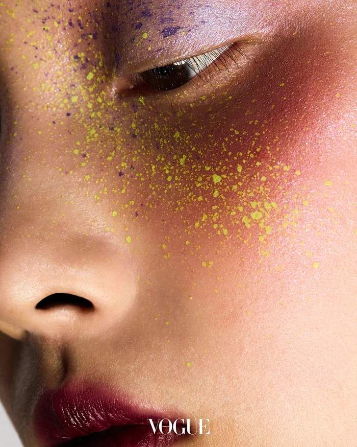 Extreme close-up beauty portrait showing an eye with purple shadow, yellow speckles across the cheek, and deep plum lipstick.