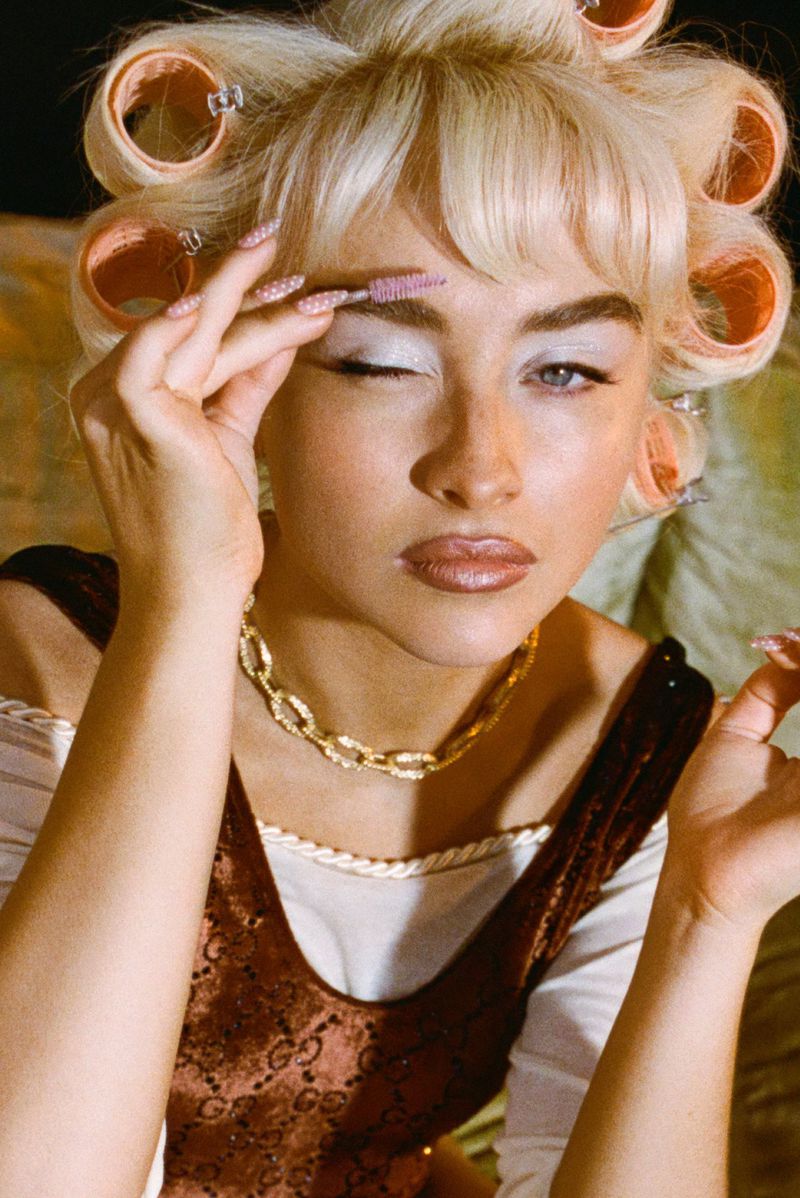 A fair-skinned woman with blonde hair in hair rollers applies makeup, wearing a brown top and gold necklace in a portrait shot.