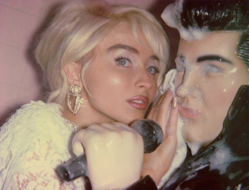 Portrait of woman with blonde hair and large earrings posing next to Elvis Presley statue covered in soap suds in front of a pink wall.