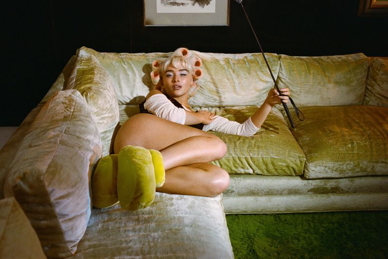 Woman lounges on a green velvet sofa with curlers in her hair, wearing a top, shorts, and slippers while holding a riding crop.