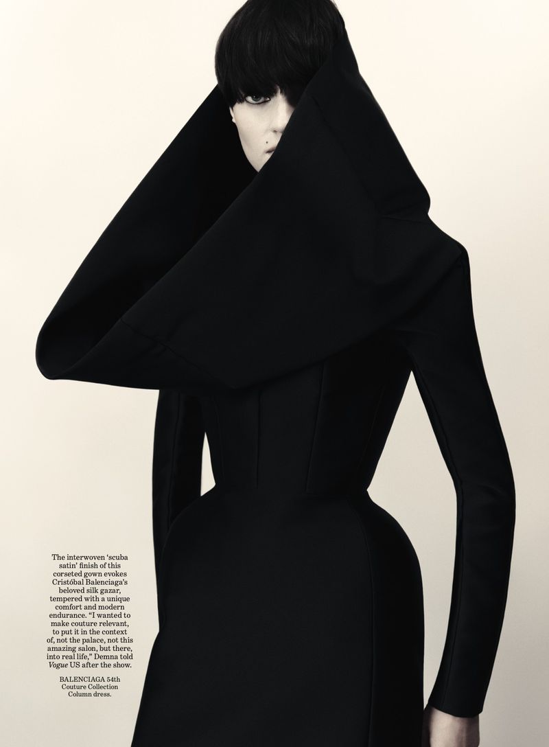 Fashion editorial shot of a woman wearing an elegant black Balenciaga gown with dramatic hooded collar and sleek, dark hair, indoor studio.